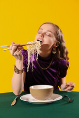 Young woman eating Chinese noodles at table on yellow background