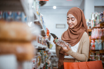 woman in headscarf holding a cell phone and a drink bottle while shopping in a store