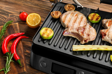 Modern electric grill with steaks and vegetables on wooden background, closeup