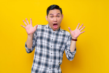 Surprised excited young Asian man wearing plaid shirt looking camera keeping his mouth wide open...