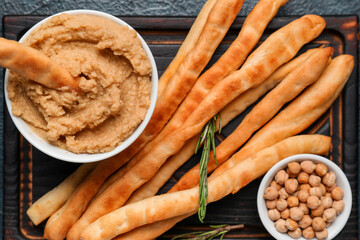 Wooden board with tasty Italian Grissini, hummus and chickpeas on dark background, closeup