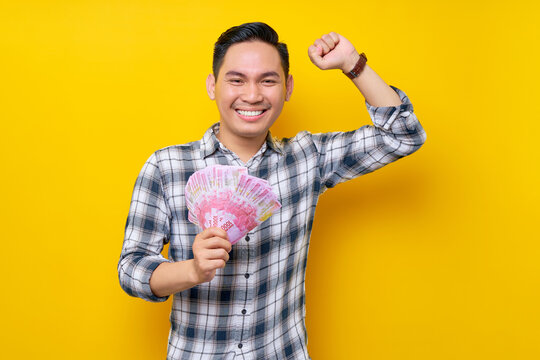 Portrait of excited young Asian man wearing plaid shirt holding cash money in rupiah banknotes and showing winner gesture isolated on yellow background. people lifestyle concept