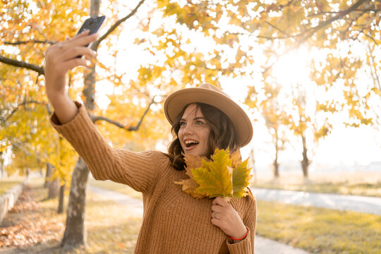 Beautiful young woman with autumn leaves taking selfie using smartphone. Season, technology and people concept.