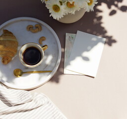 Card mockup in  breakfast still life scene with copy space.Cup of coffee, croissant , daisy flowers in a vase top view on beige desk with sunlight shadows. Feminine lifestyle composition. 