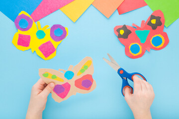 Little child hands holding scissors and cutting colorful butterfly shape from paper on light blue...