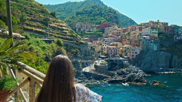 Female tourist walking in Riomaggiore harbor looking at quaint village overhanging cliffs. Cinque Terre with a small harbor beach and a wharf framed by colorful tower houses in Italy.