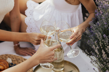 A group of girlfriends raise a toast with glasses of white colored wine on a picnic in lavender field.