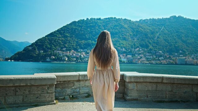 Beautiful Girl with long hair in a dress walking in slow motion by Lake Como. A female tourist approaches the edge of a viewpoint and enjoys the panoramic view of colourful houses in Italy in summer.