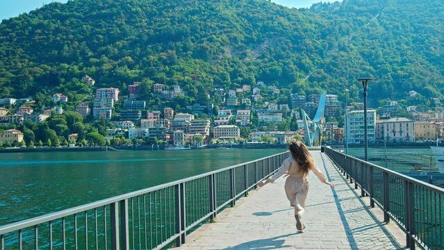Beautiful girl running in slow motion on the bridge of Lake Como in summer Italy. A young female enjoying freedom by extending her arms while running with village colourful houses in the background