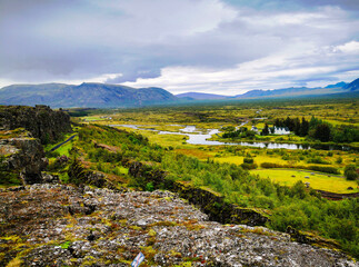 Obraz premium A stunning view of Þingvellir National Park in Iceland, featuring a lush green valley with rivers, rocky cliffs, and distant mountains under a cloudy sky