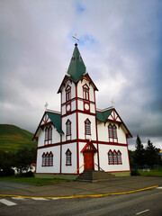 Fototapeta premium A picturesque white and red church in Iceland, standing tall with a green roof against a cloudy sky, surrounded by greenery and mountains in the background