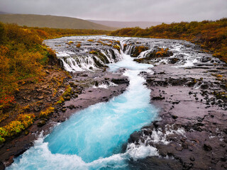 A stunning blue river flows over rocky terrain in Iceland, creating multiple cascades and surrounded by autumn-colored vegetation under a cloudy sky