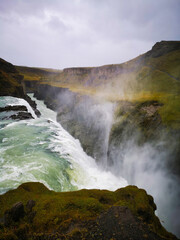 dramatic Icelandic waterfall plunges into a misty canyon, with powerful rapids flowing through rugged terrain under a cloudy sky