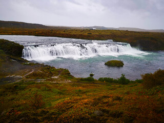 A wide, serene waterfall in iceland flows into a calm river, surrounded by a grassy landscape under an overcast sky