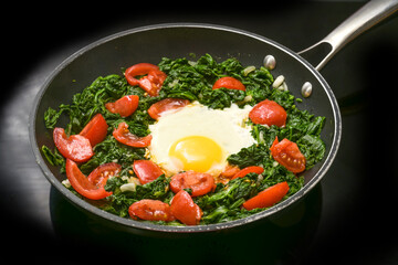 Fried egg with spinach and tomatoes in a frying pan on a black cooker top, preparing a healthy meal for breakfast or lunch, copy space, selected focus