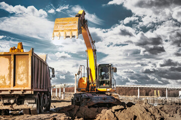 A wheeled excavator loads a dump truck with soil and sand. An excavator with a high-raised bucket...