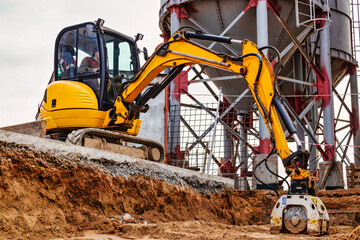 A mini excavator rams the ground with a vibrating plate. Laying of underground sewer pipes and communications during construction. soil compaction. Earthworks, excavation. © Anoo
