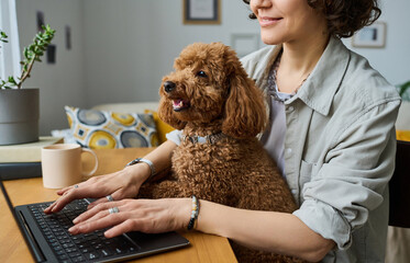 Close-up of young owner sitting at table with her dog and typing on laptop doing her online work
