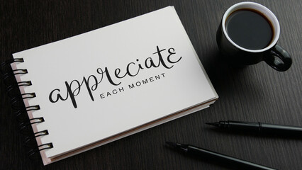 APPRECIATE EACH MOMENT black lettering in notebook with cup of coffee and pens on black wooden desk
