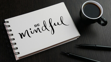 BE MINDFUL black lettering in notebook with cup of coffee and pens on black wooden desk