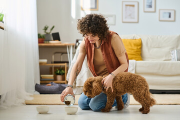 Young woman pouring food into bowl and feeding her dog in the living room at home - Powered by Adobe