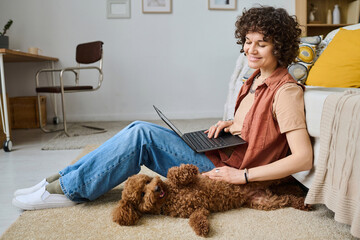 Happy young woman sitting on the floor in the living room with laptop on her knees and playing with her dog