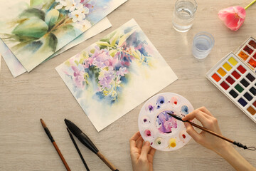 Fototapeta na wymiar Woman painting flowers with watercolor at white wooden table, top view. Creative artwork