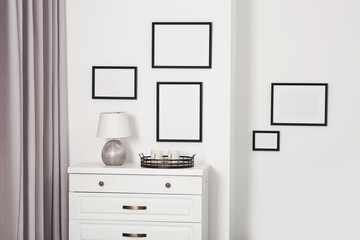 Empty frames hanging on white wall, wooden chest of drawers and lamp indoors