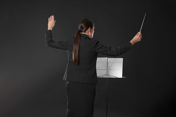 Professional conductor with baton and note stand on dark background, back view
