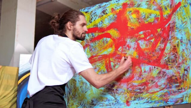 Innovative talented artist with long hair, wearing white t-shirt, creates masterpiece using red and blue colors. Man paints with brush on a large canvas in creative studio on background of paintings.