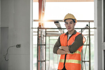 Portrait of asian man architect at building site with folded arms looking at camera. Confident construction manager in formal clothing wearing yellow hardhat. Successful engineer