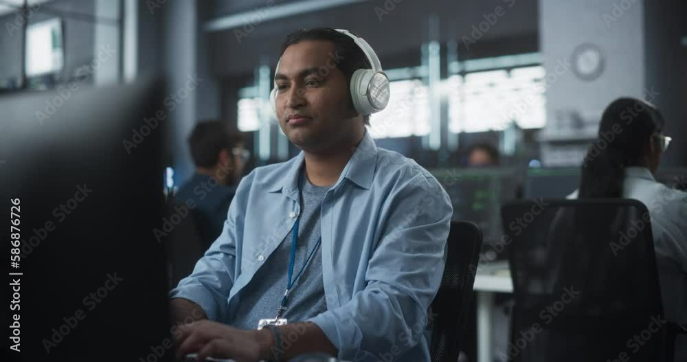 Poster Indian System Administrator Working in a Research E-Business Facility on a Desktop Computer. Focused Software Developer in Casual Clothes Wearing Headphones and Updating Server System Database