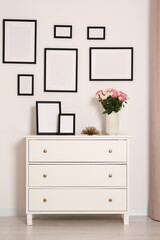 Empty frames hanging on white wall and chest of drawers with flowers in room