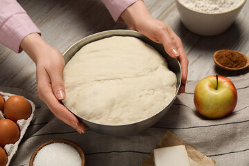 Woman holding bowl with fresh yeast dough and ingredients for cake on wooden table, closeup