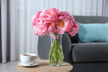 Beautiful bouquet of pink peonies in vase and cup with drink on wooden table indoors