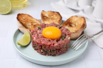 Tasty beef steak tartare served with yolk, toasted bread and lime on white tiled table