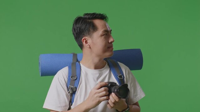 Close Up Side View Of Asian Male Hiker With Mountaineering Backpack Using A Camera Taking Picture While Standing On Green Screen Background In The Studio

