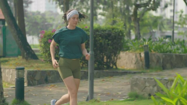 Slow motion shot of young fit woman in sportswear running along sidewalk in park while training outdoors in morning