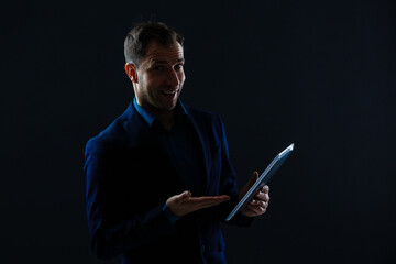 A serious handsome young businessman in a suit using a tablet in front of a dark grey background in a studio.