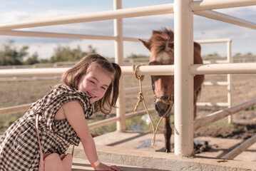 A cute little girl leaning on a feeding pen looking at the camera, behind her is a horse in a stable.