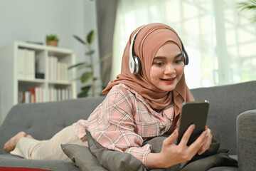 Obraz premium Smiling beautiful Muslim woman wearing hijab and smart casual wears lying on couch and using mobile phone