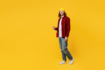 Fototapeta na wymiar Full body sideways smiling happy fun young man wearing red checkered shirt white t-shirt hat walking going strolling look camera isolated on plain yellow background studio portrait. Lifestyle concept.