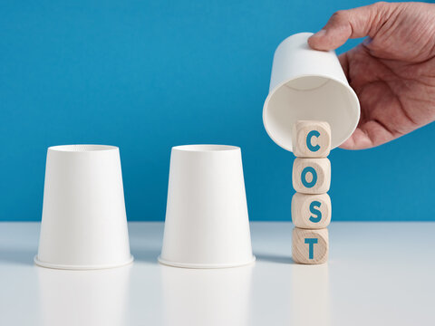 Finding the hidden costs concept. Man playing the shell game with three cups reveals the word cost on wooden cubes.