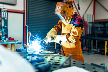 A woman in protective equipment uses tools and machines in the workshop, sparks fly and illuminate the workshop