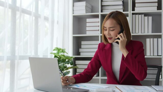 Beautiful Asian businesswoman working on a laptop computer using a mobile phone to negotiate financial business accounting analysis, report, and office information.