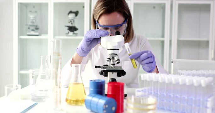 Chemist holds test tube with oil and looks through microscope in research laboratory. Testing concept of oil properties