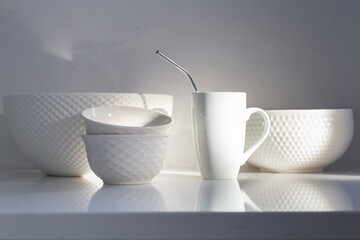 Selective focus view of white ceramic bowls and coffee cup with metal straw set on quartz countertop in rays of sunlight