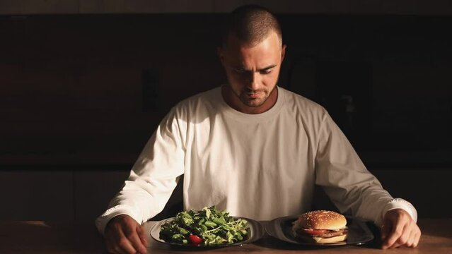 Man make choice salad or burger, unhealthy or healthy food, diet. Male look with aversion on salad and choose burger pushed the plate away. Male make decision eat unhealthy food, show thumb up
