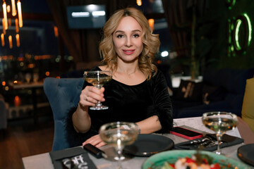 woman in a restaurant with champagne in her hand. She is sitting at a table against the backdrop of a dark restaurant hall with lamps.