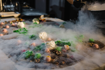 Pastry chef wearing black uniform holding a bowl preparing delicious chocolate at kitchen. Cooking chocolate. Hands pouring cooled liquid nitrogen on hot chocolate. Chocolatier cooling melted mass.